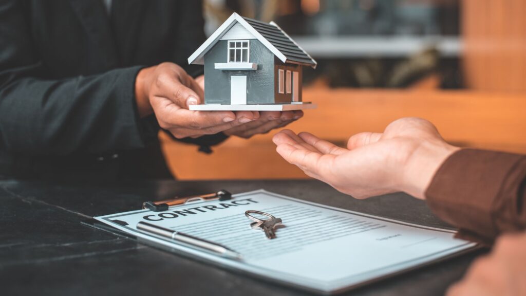 A person in a suit hands a small house model to another, with a contract and keys on the table, symbolizing a real estate transaction.
