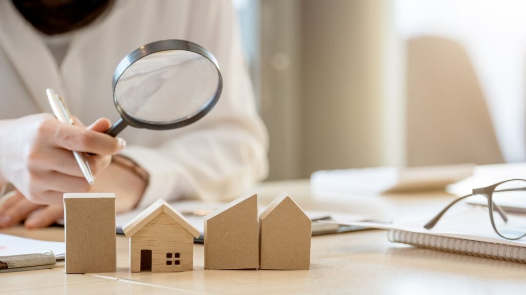 Hand holding a magnifying glass over wooden house models on a desk, symbolizing property evaluation.