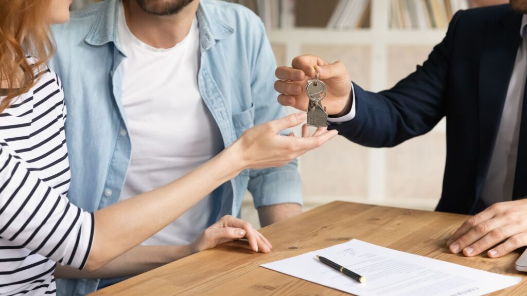 A person handing over house keys to a couple at a table with documents, symbolizing a property transaction.