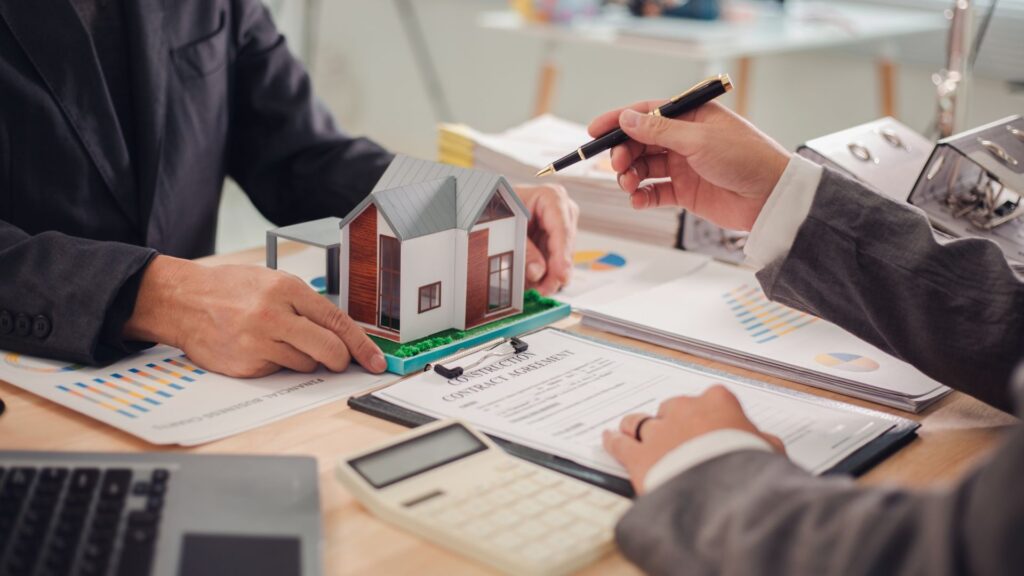 Two people in suits reviewing a house model and construction agreement with charts and a calculator on the table.